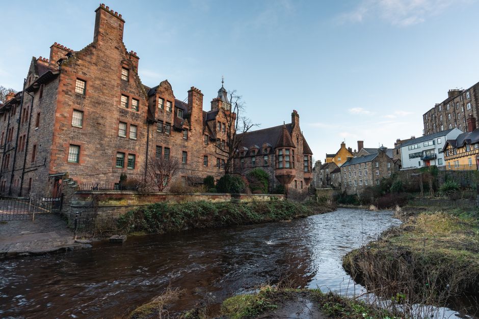 The image shows a view of a residential area along a small river or stream, with historic stone and brick houses built closely together on both sides of the water. Some of the buildings have multiple