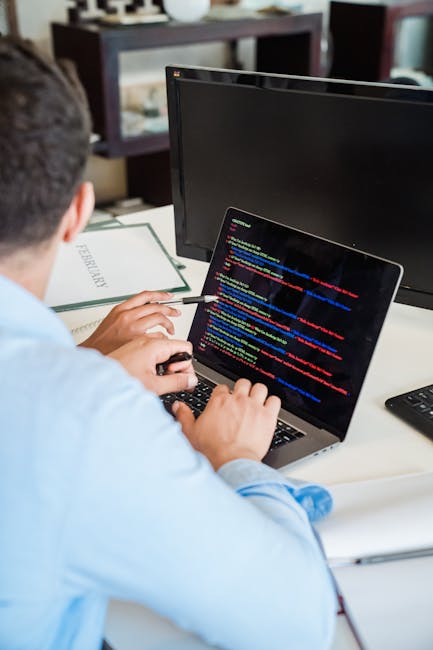 A person wearing a light blue shirt sits at a desk inside a home or office, working on a laptop that displays lines of colourful computer code on its screen. The workspace includes a larger external m